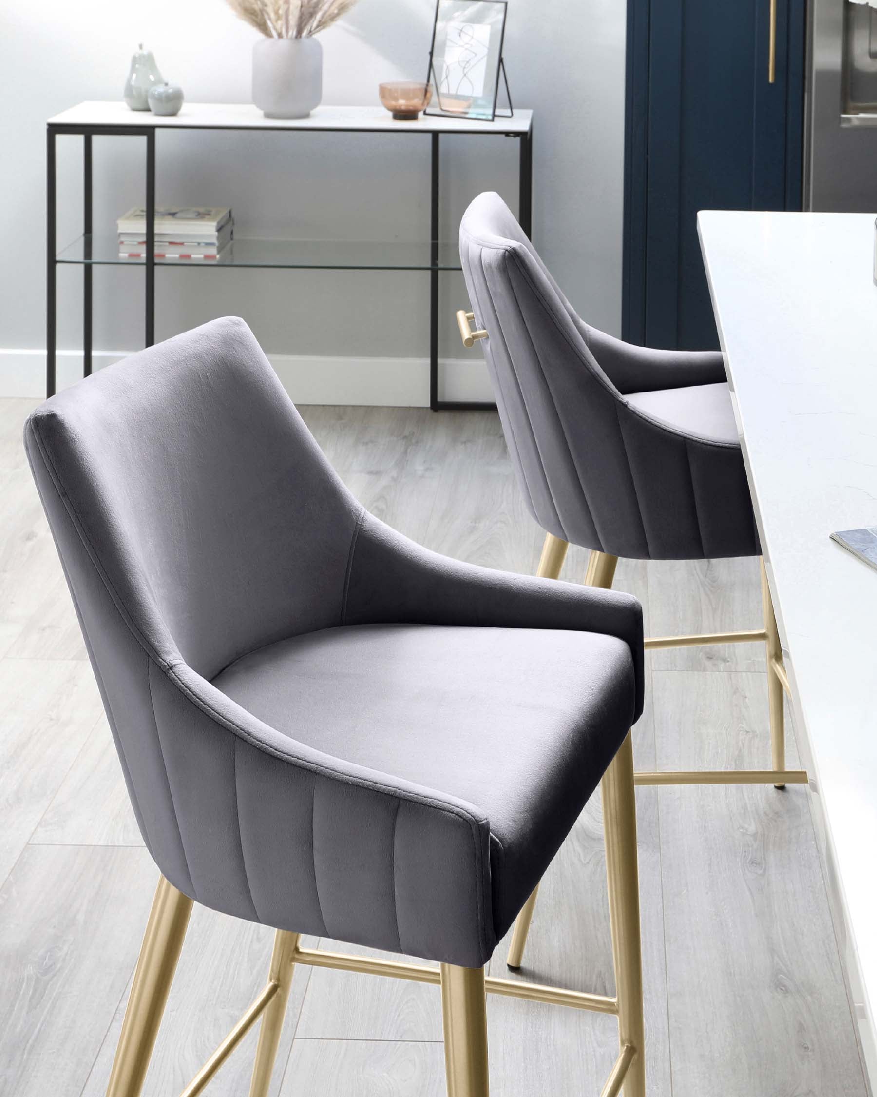 Elegant modern bar chairs with grey upholstery and gold metal legs, positioned around a white marble-top counter. A minimalist console table with black metal frame and glass shelves, adorned with decorative items, is visible in the background.