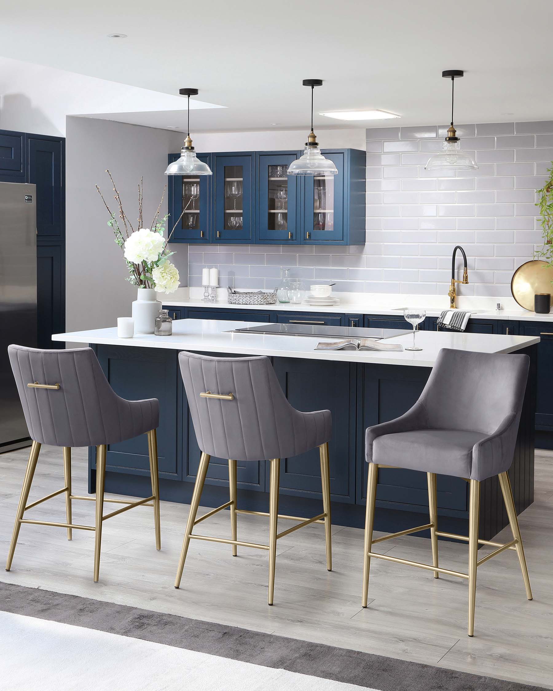 Three contemporary bar stools with upholstered seats in grey fabric and brass-finished metal legs, featured at a kitchen island.