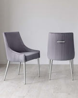 A pair of modern grey upholstered chairs with sleek, silver metal legs. The chair on the left features a curved backrest and comfortable armless seat design, while the chair on the right has a high back with distinctive vertical stitching and a decorative silver handle at the top. Both are positioned on a light wooden floor against a neutral background.