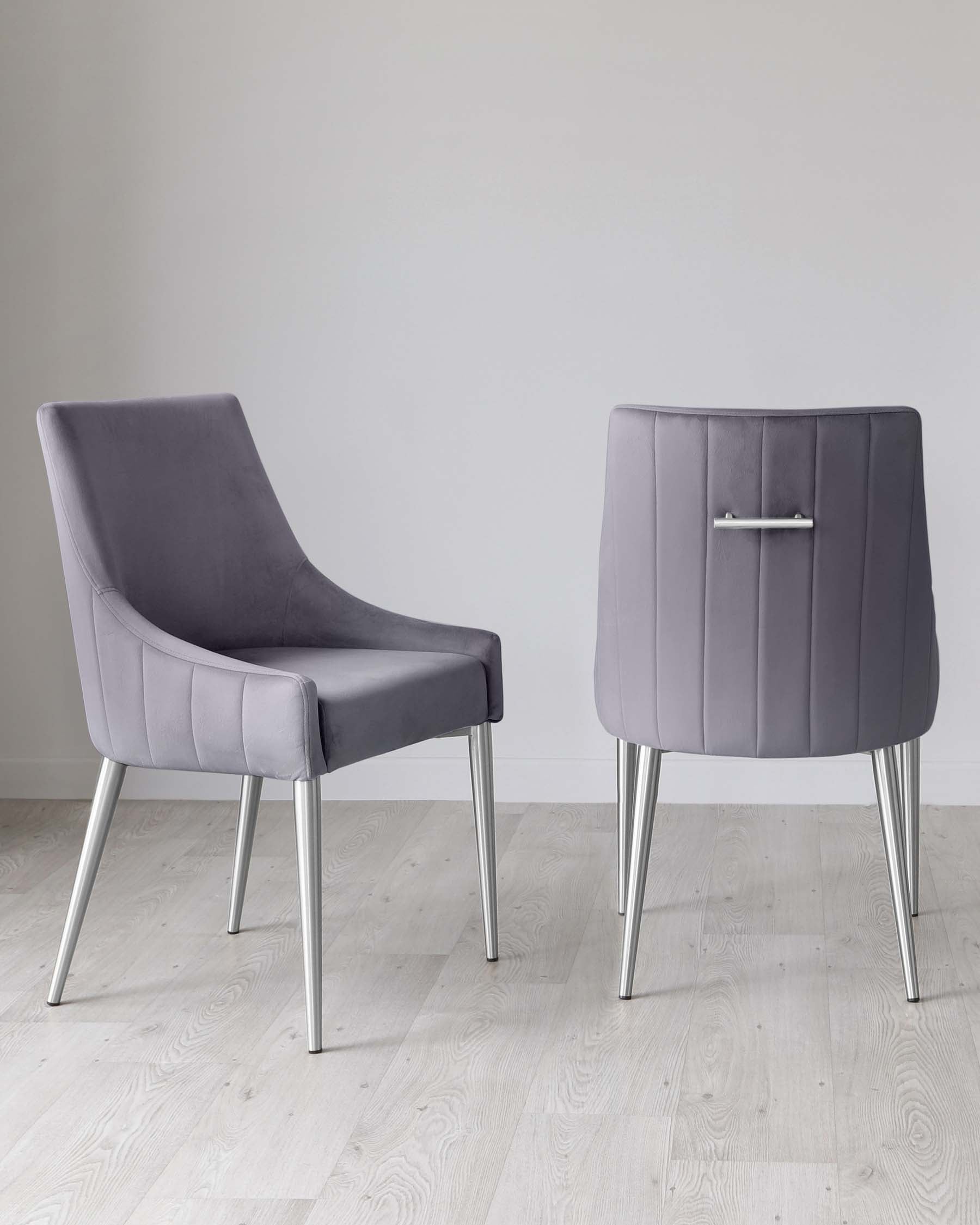 A pair of modern grey upholstered chairs with sleek, silver metal legs. The chair on the left features a curved backrest and comfortable armless seat design, while the chair on the right has a high back with distinctive vertical stitching and a decorative silver handle at the top. Both are positioned on a light wooden floor against a neutral background.