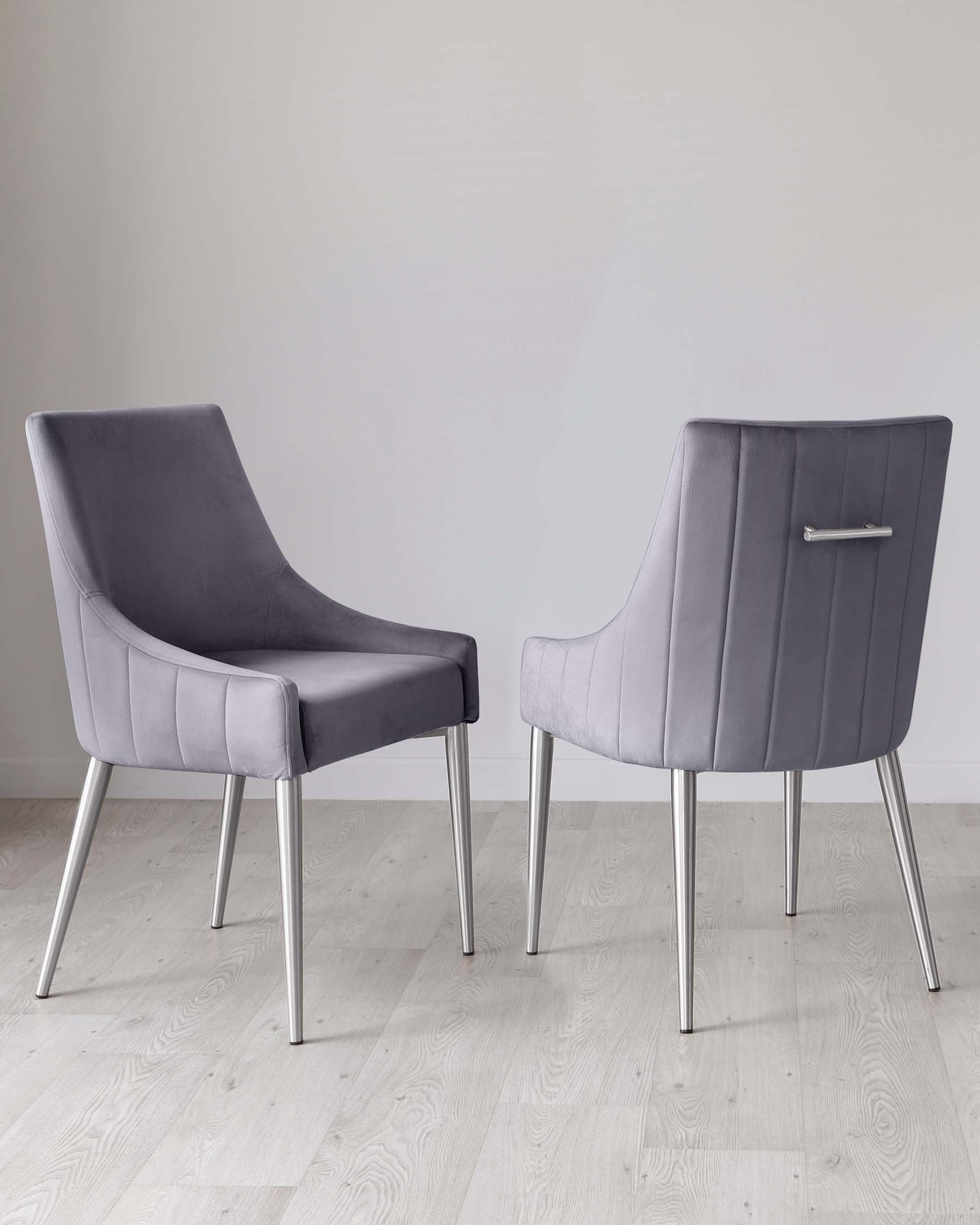 Two modern dining chairs with sleek grey upholstery and horizontal channel tufting. The chairs have slightly curved backrests and are supported by polished chrome legs. The design presents a blend of comfort and contemporary elegance, displayed on a light hardwood floor against a white wall background.