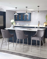 Modern kitchen with three stylish upholstered bar stools in grey fabric with channel tufting and brushed stainless steel legs, positioned at a white island counter.