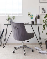 Contemporary office setup featuring a stylish, ribbed grey upholstered desk chair with a high backrest and armrests, mounted on a polished silver five-point wheeled base. The chair is paired with a minimalist glass top desk supported by sleek, black A-frame metal legs. The scene is complemented by modern decorative elements including plants and books, suggesting a clean and productive workspace.