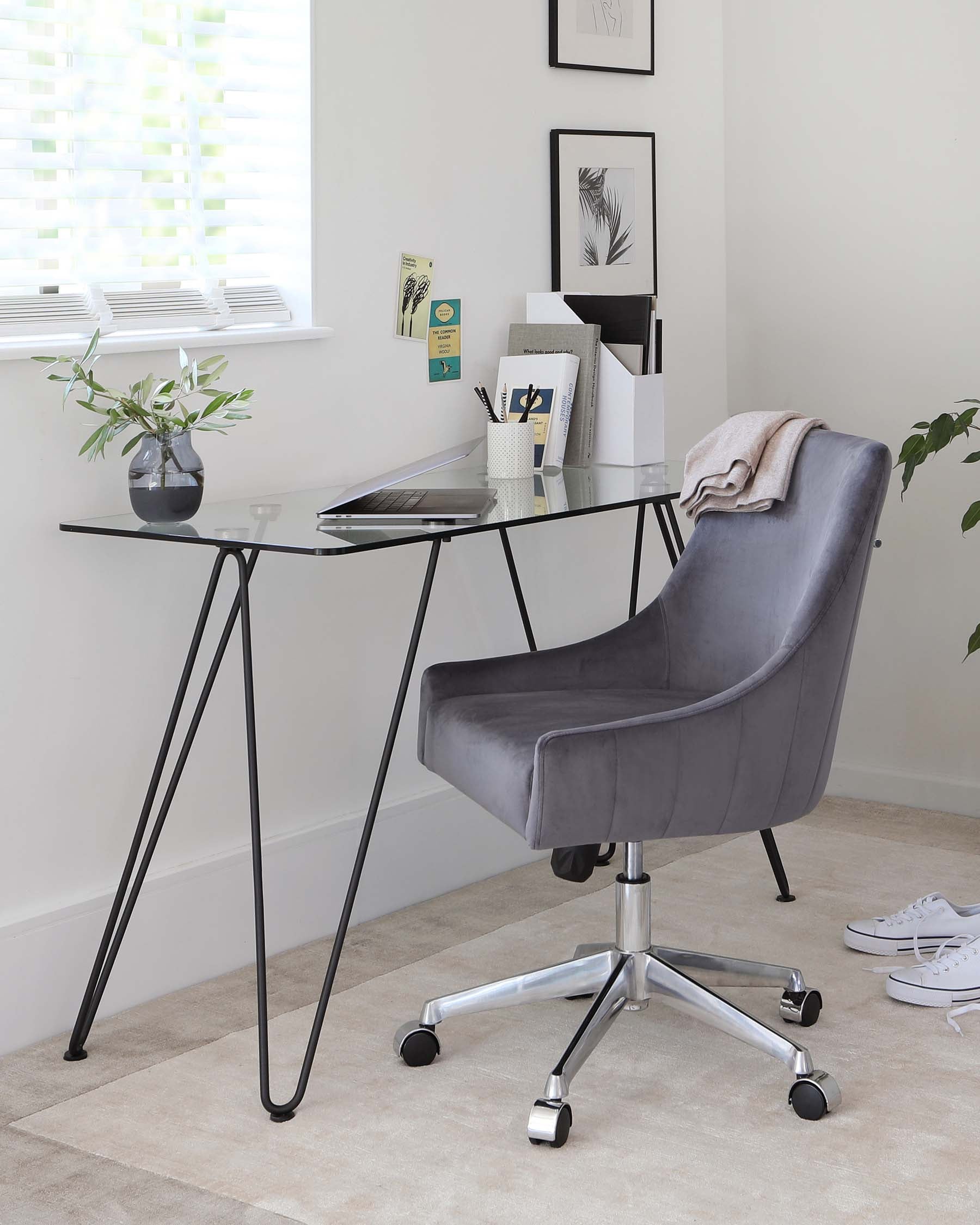 Modern minimalist office setup featuring a sleek glass-top desk with black metal hairpin legs and a comfortable grey upholstered swivel chair with a high backrest and shiny chrome base with rolling casters.