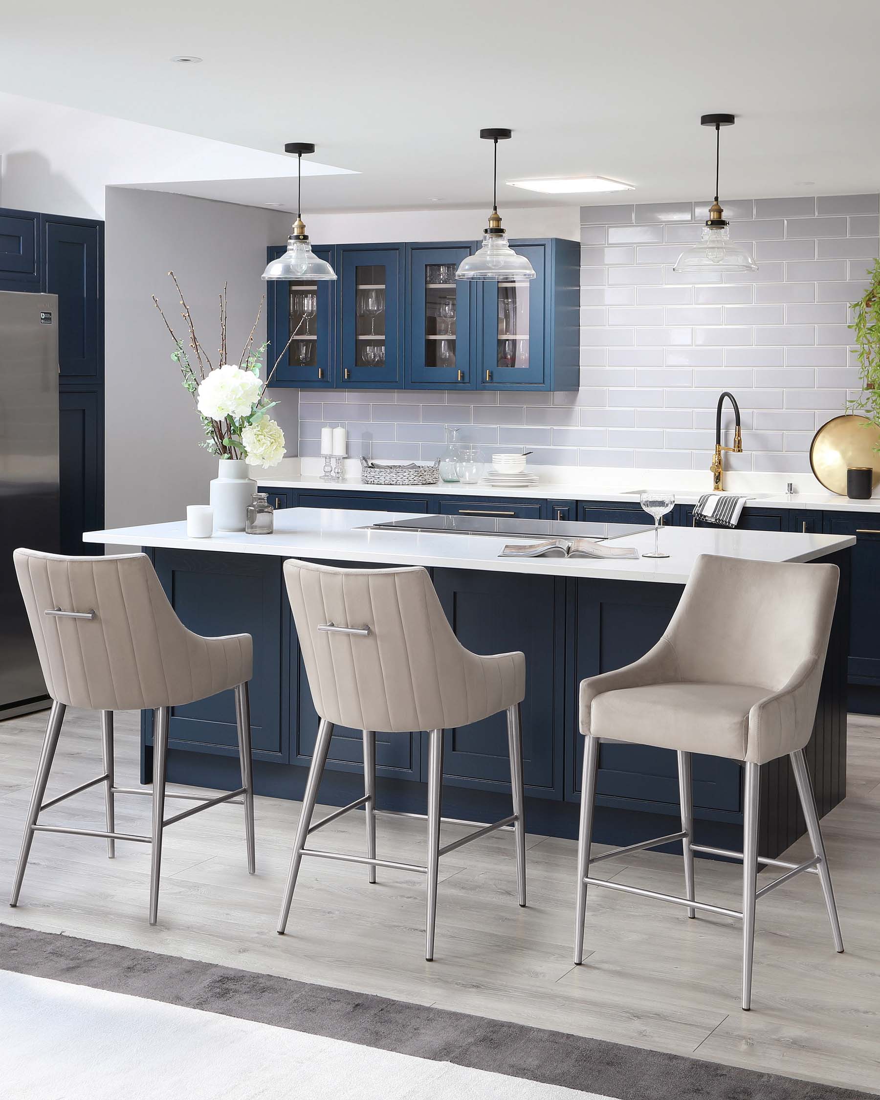 Three modern light grey upholstered bar chairs with high backs and metal legs are set against a kitchen island with a white countertop and navy cabinetry.