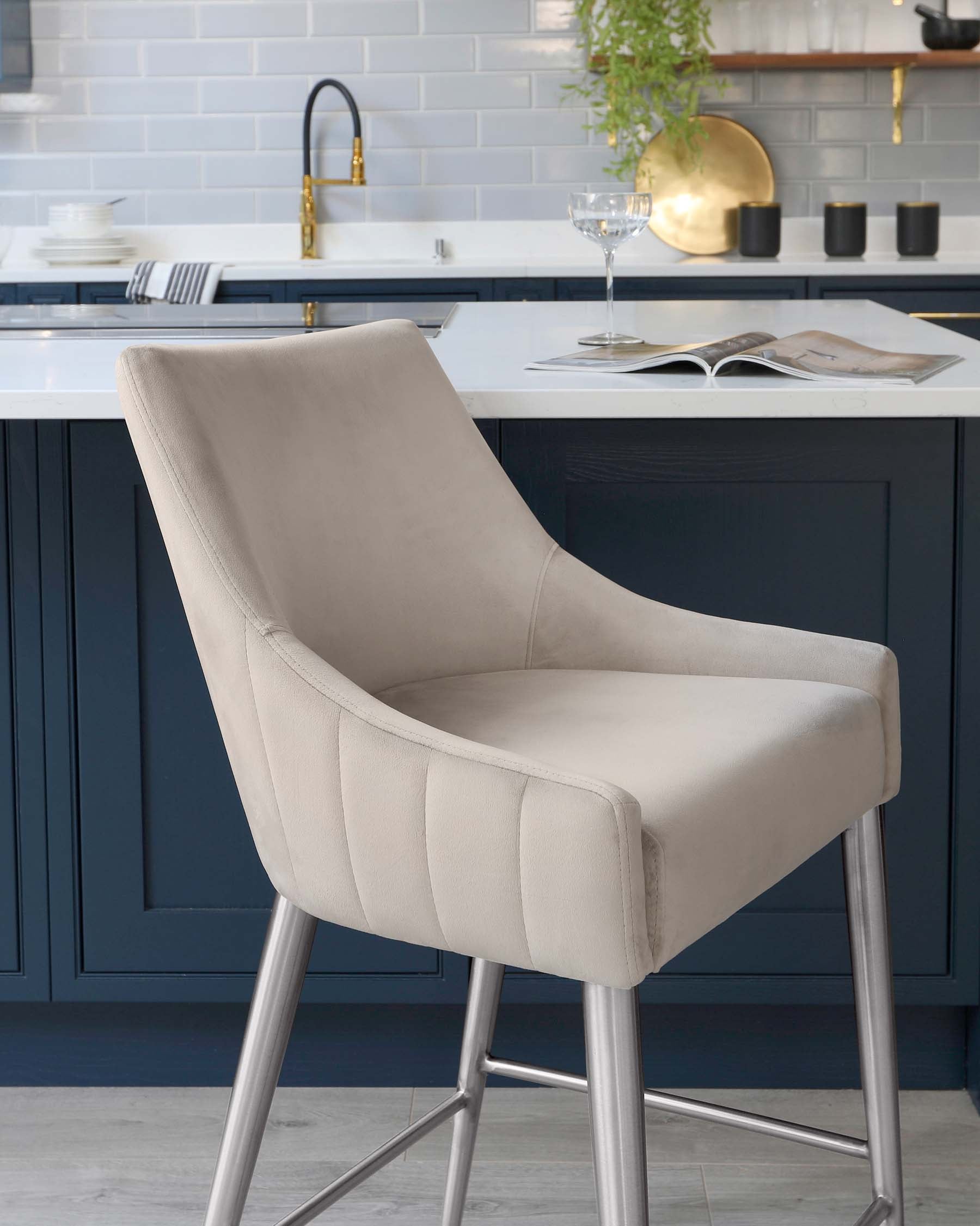 Elegant modern bar stool with a sleek, light-coloured fabric upholstery and horizontal stitching details on the backrest. The stool has a comfortable curved design and is supported by slender, metallic legs that give it a contemporary and chic look. The background features a stylish kitchen setting with blue cabinets and white countertops.