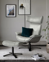 Modern light grey leather lounge chair with a high backrest and matching ottoman, featuring metal swivel bases. A dark teal throw pillow accents the chair. A sleek floor lamp with a glass shade stands nearby. Magazines are casually placed on the floor next to a black coffee cup. The furniture is arranged on a textured grey rug in a minimalist styled room.