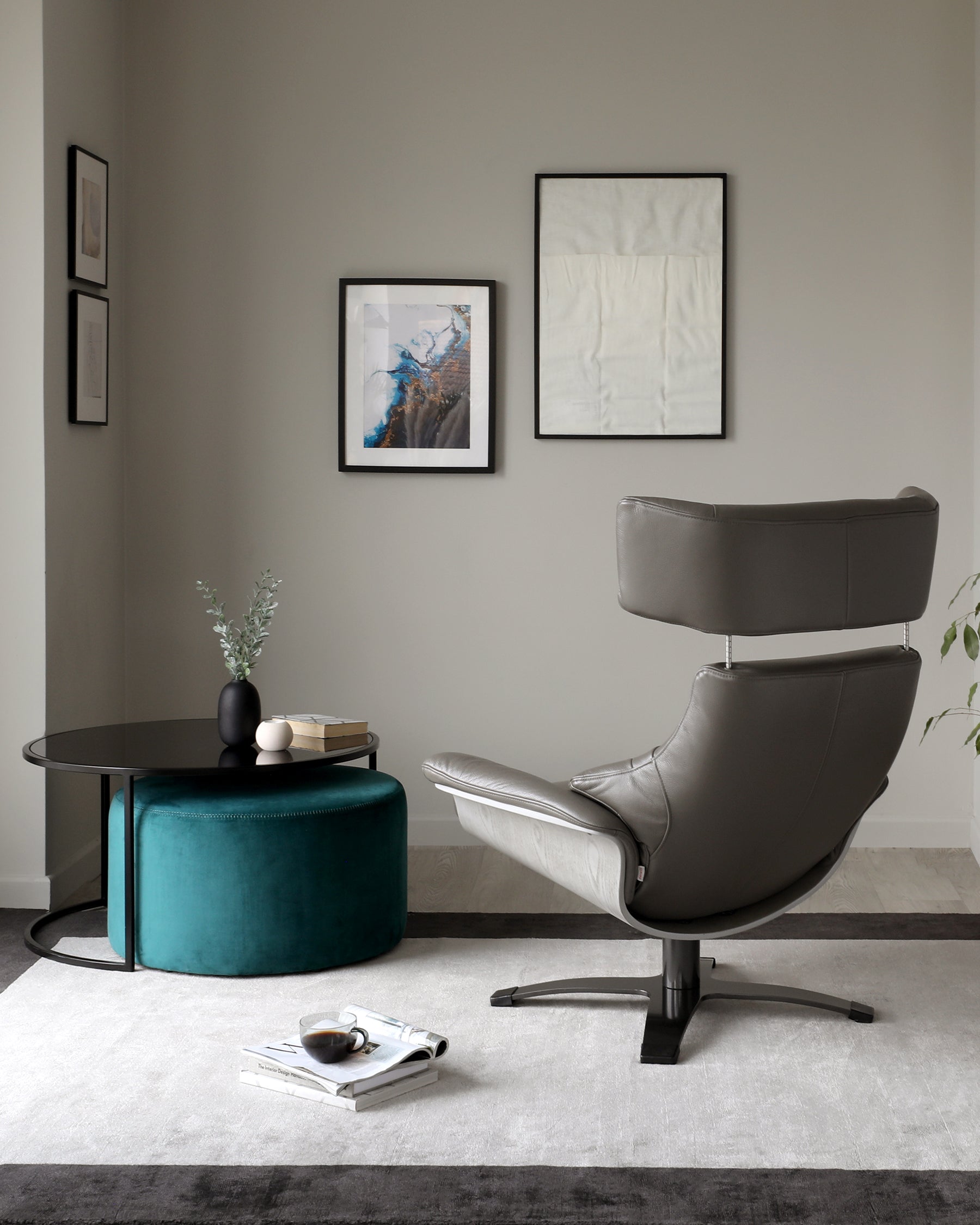 Contemporary room with a matte black round coffee table alongside a teal upholstered ottoman, and a sleek grey leather lounge chair with a high backrest and metal base. A light grey area rug underneath lays on a dark hardwood floor.