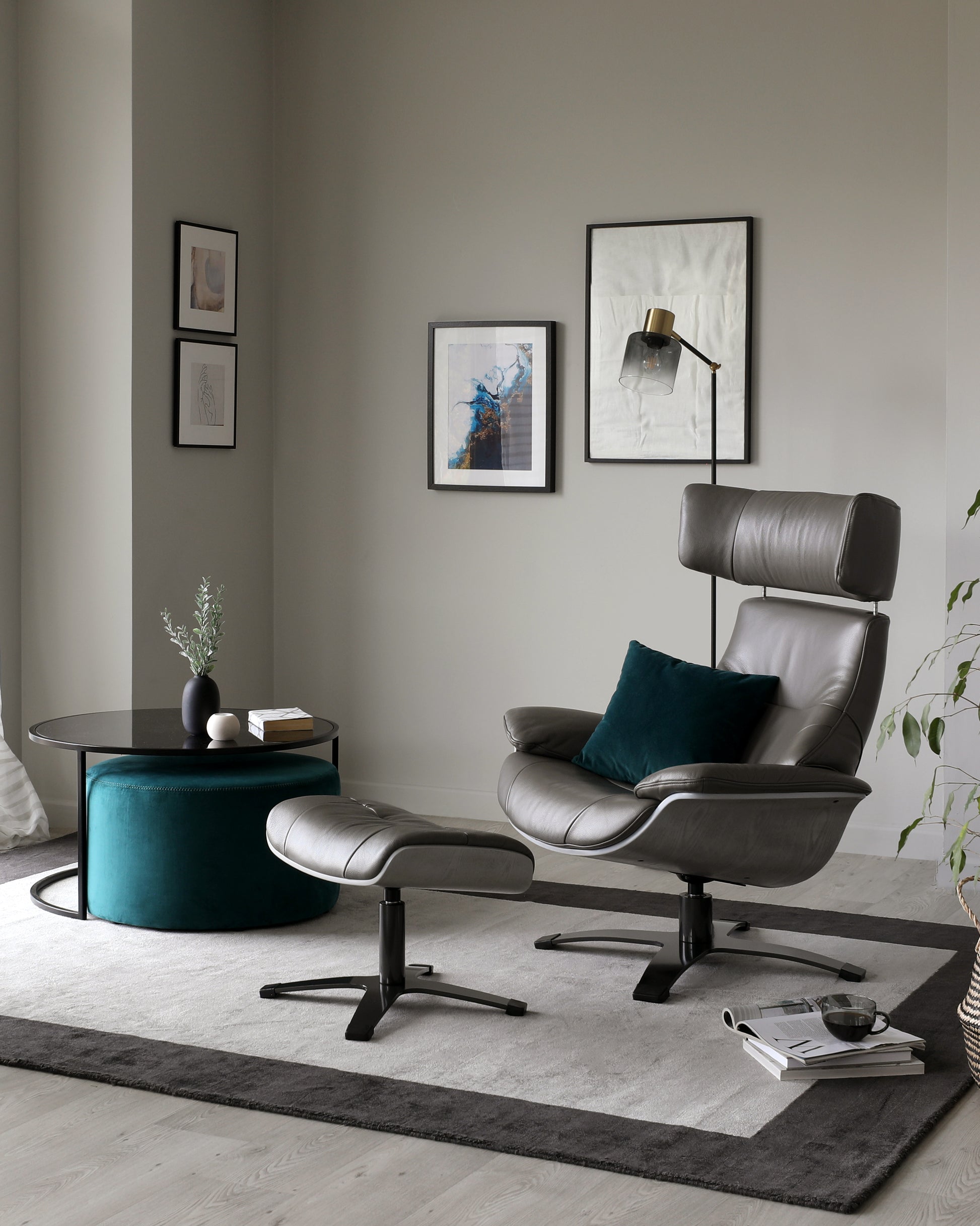 Modern minimalist living room interior with a dark-toned, round coffee table and a matching footstool, complemented by a sleek, grey upholstered recliner chair with a padded headrest and matching ottoman, accented with a dark green decorative cushion. The ensemble is arranged on a grey area rug with dark border detailing.