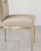 Elegant modern chair with a beige upholstered seat and a curved backrest on sleek golden metal legs set against a light hardwood floor and white wall.