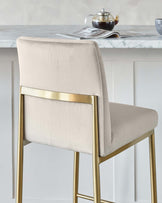 Elegant modern bar stool with a beige upholstered seat and backrest, and sleek gold-finished metal frame with a footrest.