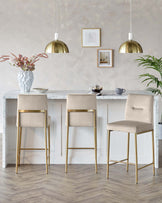 Three modern bar stools with beige upholstery and gold-finished metal legs positioned at a marble countertop, complemented by stylish pendant lights with a brass finish.