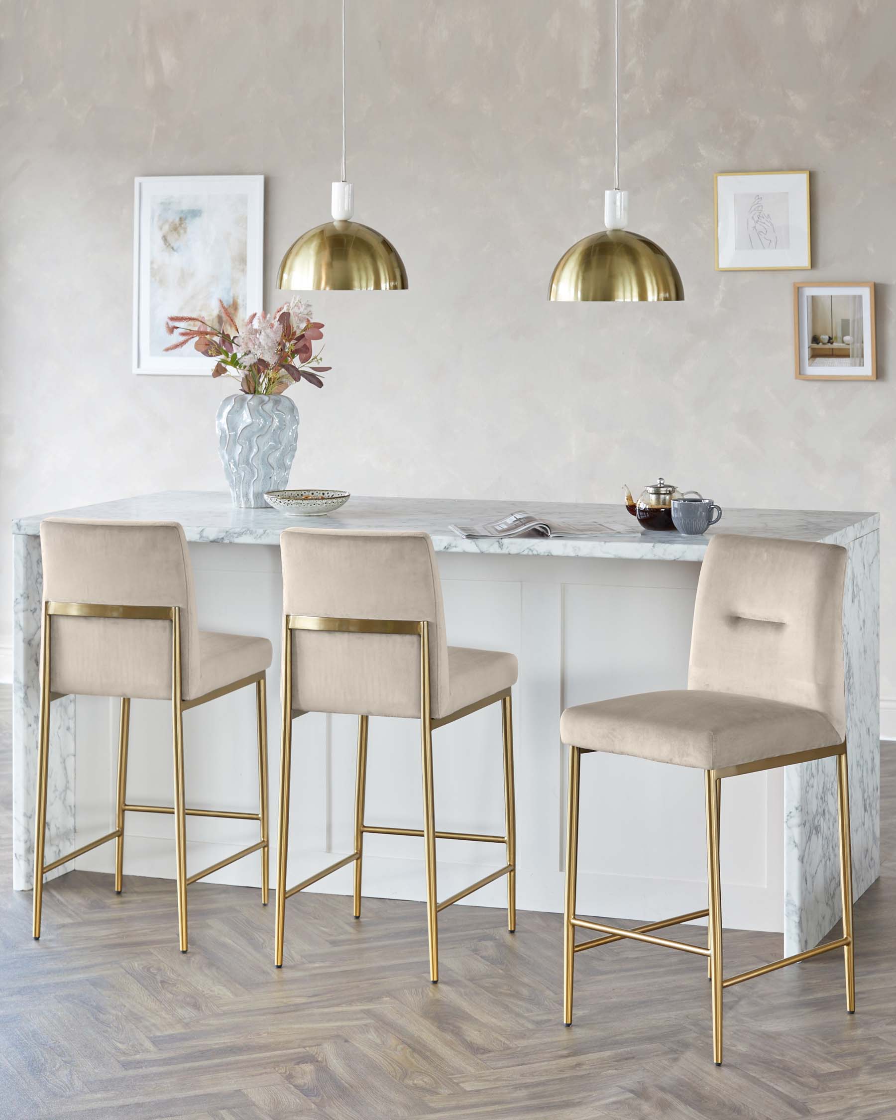 Elegant modern bar stools with beige upholstery and gold metal frames, set against a white marble countertop kitchen island under brass pendant lights.