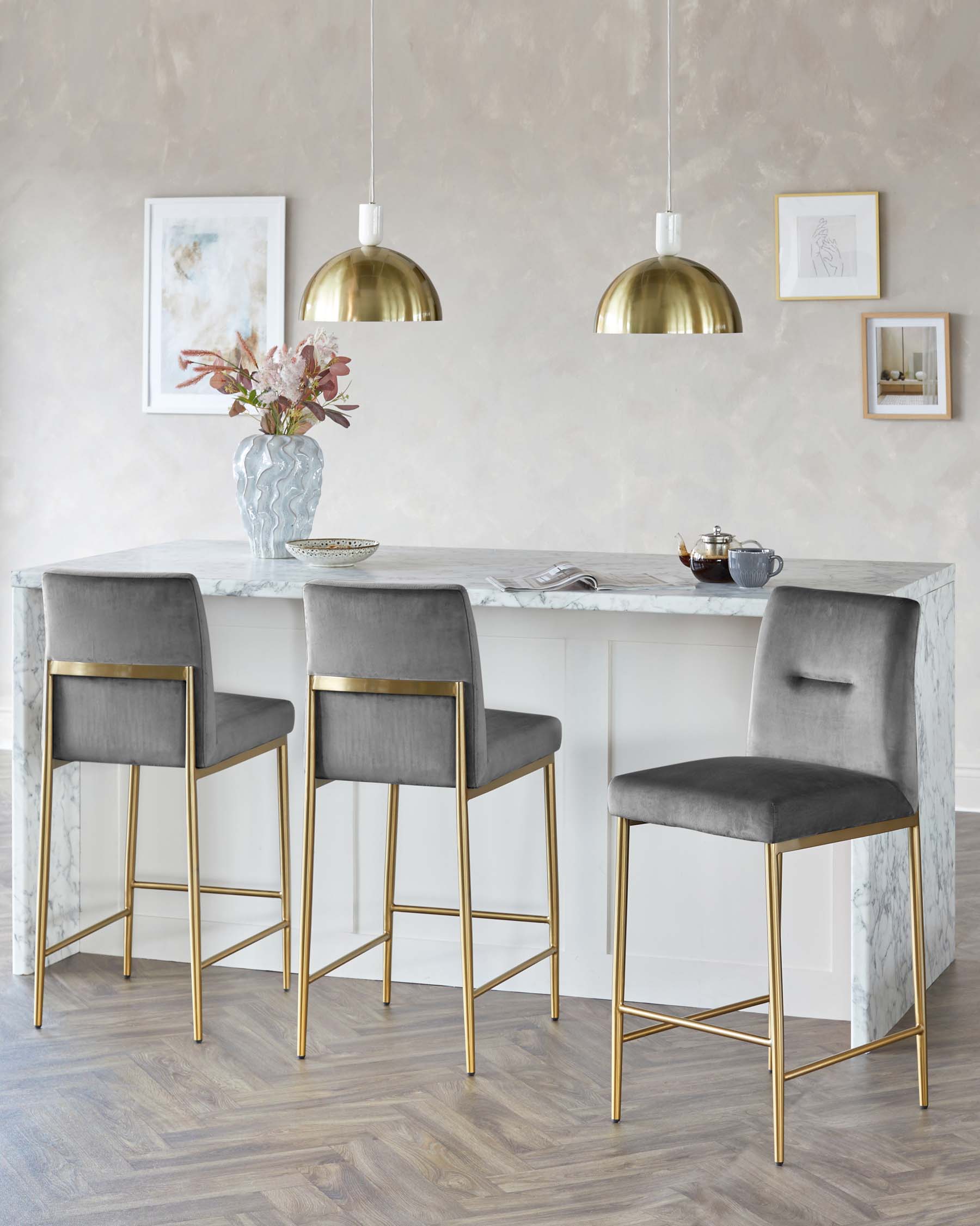 Elegant dining space featuring three grey velvet upholstered bar stools with gold metal legs, complementing the white marble countertop of the kitchen island. Additionally, the kitchen showcases two brass pendant lights with a metallic finish casting a warm ambiance.