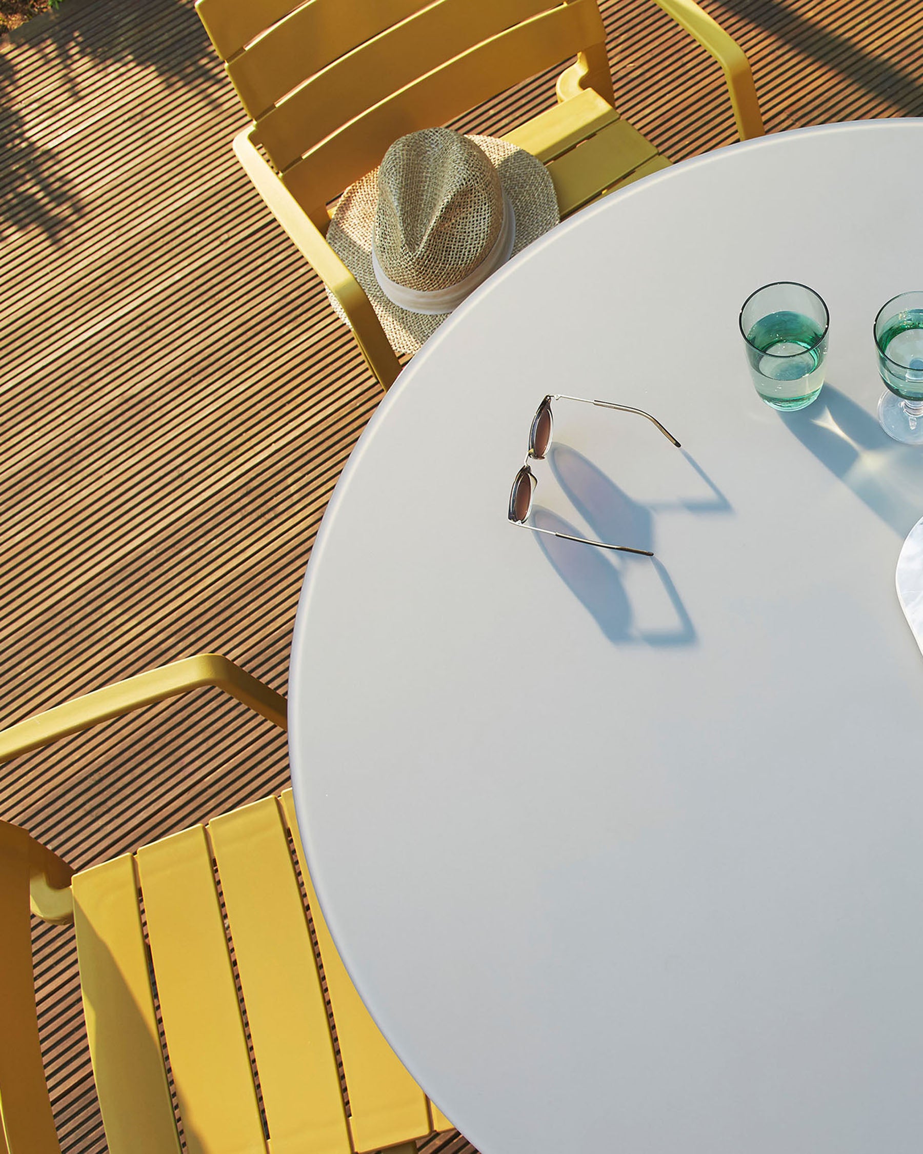 Modern outdoor furniture set featuring a round white table and a yellow slatted metal chair, on a wooden deck bathed in sunlight.