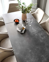 Modern dining setting featuring a large rectangular table with a dark, stone-finish surface and simple, straight edges. Accompanying the table are luxurious taupe upholstered dining chairs with a smooth, rounded design and substantial cushioning, offering comfort and elegance. The contemporary aesthetic is emphasized by the clean lines and minimalist decor.