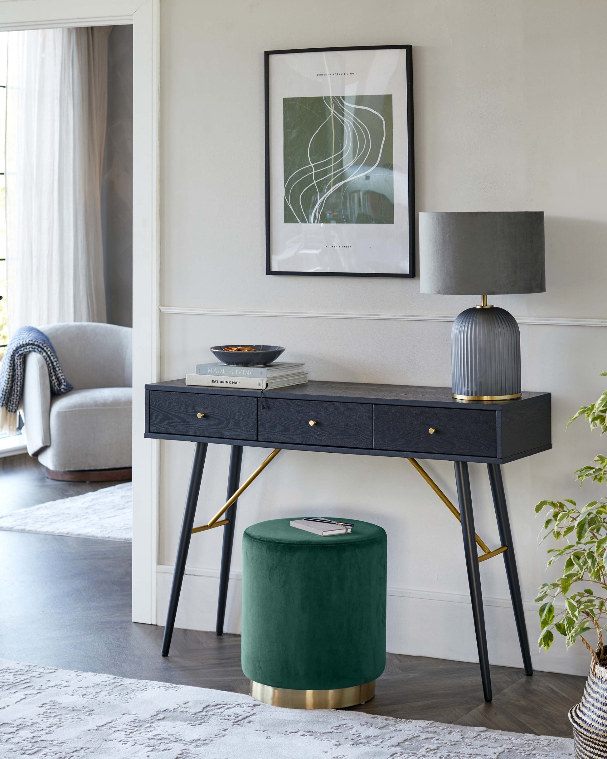 An elegant, modern console table in dark wood finish with slender, angled legs in a contrasting brass colour, featuring three drawers with minimalist pulls. Accompanied by a plush, round, green velvet ottoman with a brass base. Decor includes a grey lamp with a ridged base and a cylindrical shade, and an assortment of books atop the console.