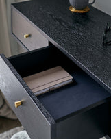 Black textured wooden nightstand with brass handles. The nightstand features an open drawer revealing a lined interior with a brown notebook inside, atop the closed upper drawer sits a cup and saucer with a pair of eyeglasses.