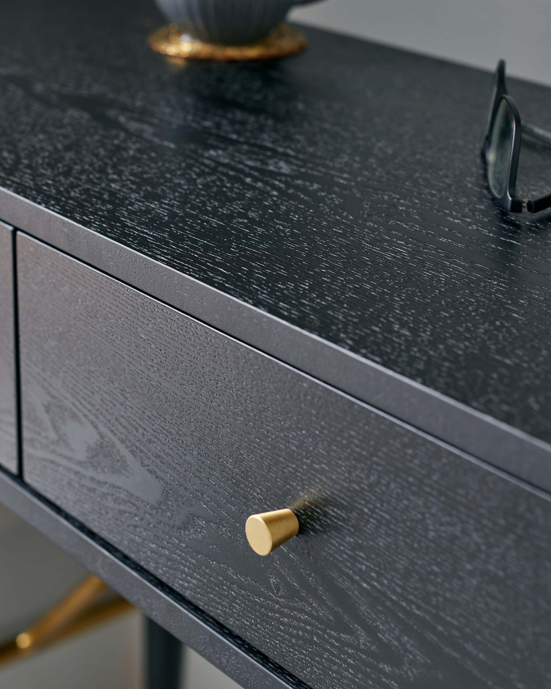 Elegant black textured wooden console table with a sleek brass knob on a drawer, featuring brass accents on leg tips and stylish accessories including a golden-rimmed bowl and fashionable glasses atop the surface.