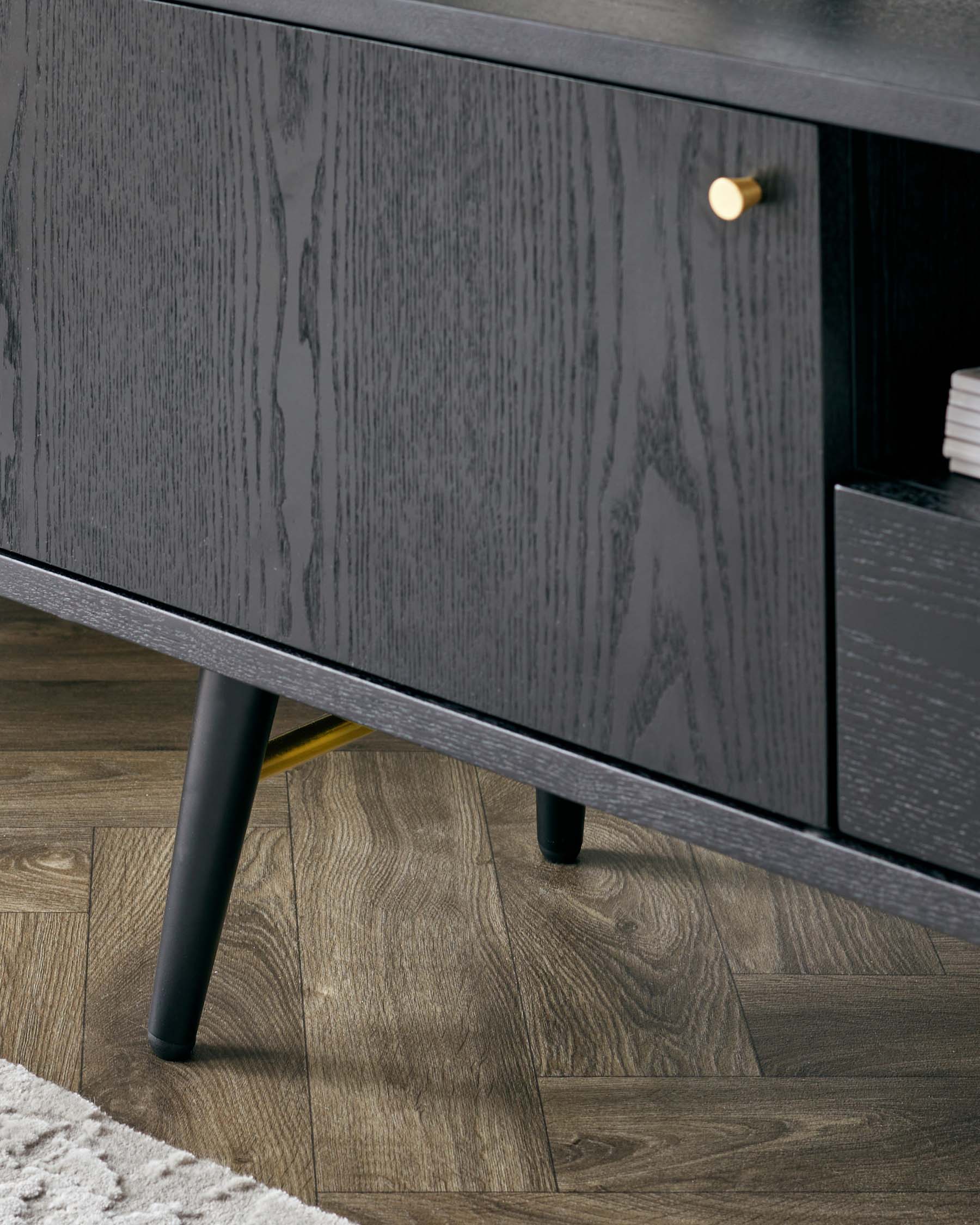 A modern dark wood sideboard with visible grain detail, featuring sleek lines and angled black metal legs. One of the drawers is partially open, displaying a single brass knob handle. The sideboard sits on a herringbone wood floor beside a white textured area rug.