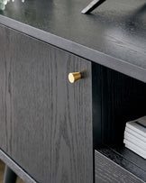 Elegant black wooden console table with a smooth top, visible grain texture, and a gold-coloured cylindrical drawer knob. There is an open shelf with a stack of white books below the drawer.