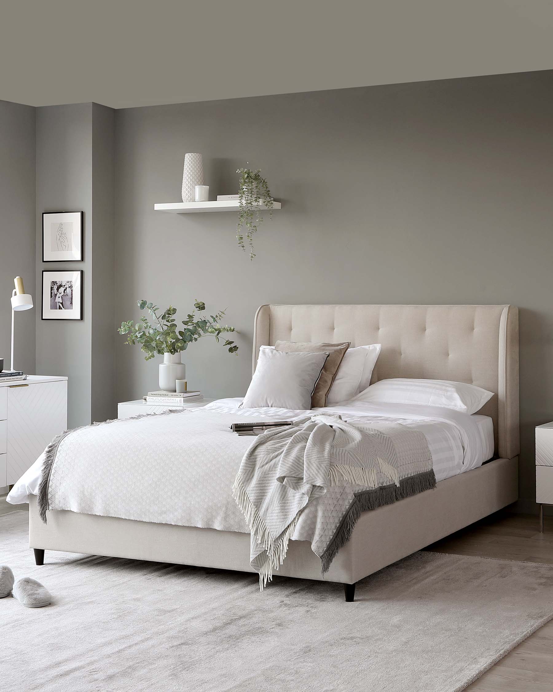 Elegant beige upholstered bed with a high tufted headboard and a neutral-toned bedding set. Beside the bed is a white modern nightstand with minimalistic design and a table lamp on top. A matching understated white floating shelf hangs above, adorned with decorative vases and greenery.