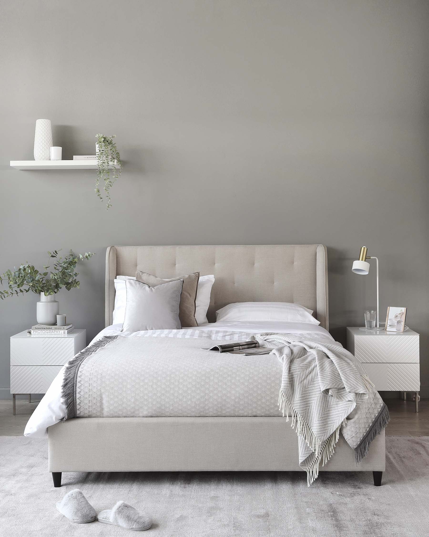 A contemporary bedroom featuring a beige upholstered bed frame with a tufted headboard, two white nightstands with a geometric facade design, and a minimalist white wall shelf.