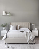 Elegant bedroom furniture set including a beige upholstered bed with a tufted headboard, two white nightstands with a geometric front design, and a white minimalist floating shelf above the bed. The scene is complemented by white and grey bedding, accent pillows, a grey throw blanket, decorative vases, and a small table lamp.