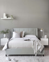 Contemporary bedroom furniture set featuring a light grey upholstered bed with a tufted headboard, two white bedside tables with a geometrical pattern design, and a white floating shelf mounted on a grey wall. The room is accessorized with a grey and white patterned bedding set, light grey throw blanket, assorted pillows, decorative vases, framed artwork, and a modern gold and white bedside lamp.