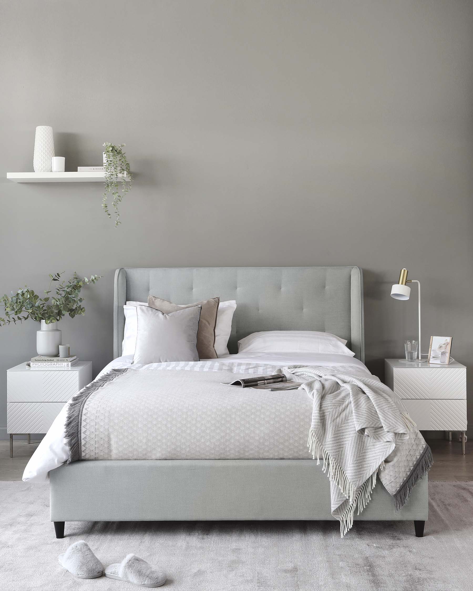 Contemporary bedroom furniture set featuring a light grey upholstered bed with a tufted headboard, two white bedside tables with a geometrical pattern design, and a white floating shelf mounted on a grey wall. The room is accessorized with a grey and white patterned bedding set, light grey throw blanket, assorted pillows, decorative vases, framed artwork, and a modern gold and white bedside lamp.