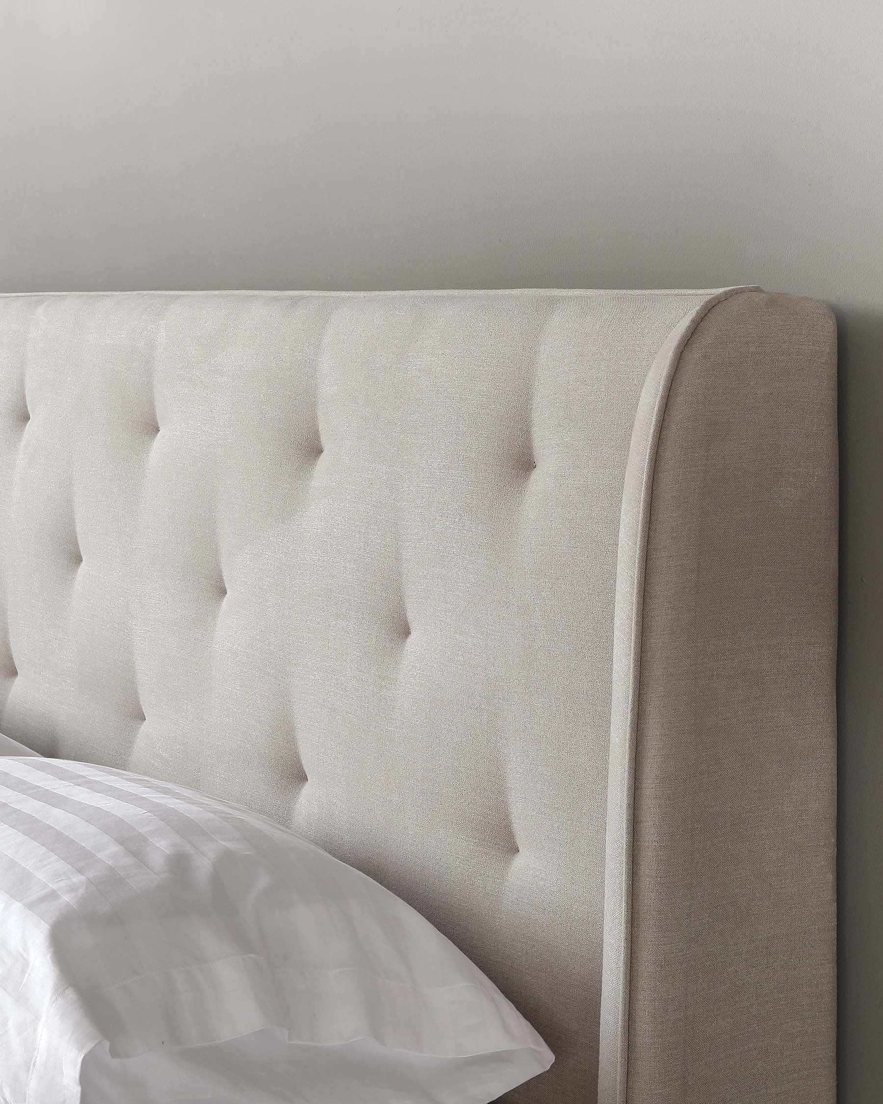 Elegant upholstered headboard featuring a tufted design in a neutral beige fabric, with a smooth top edge and side wings that frame the bed. A section of a white bedsheet is visible, indicating its use on a bed.