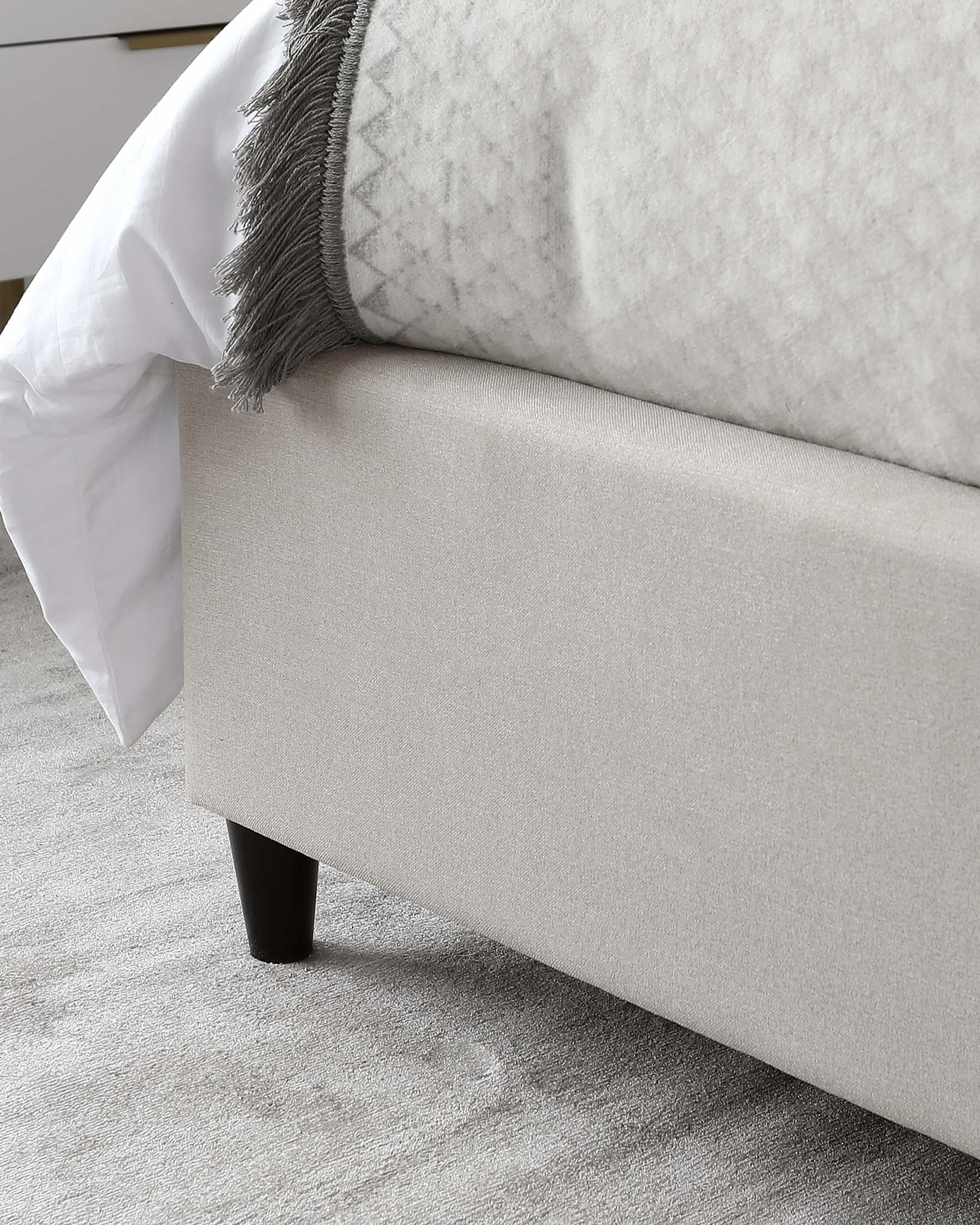 Contemporary light beige upholstered platform bed with clean lines and black tapered legs, partially dressed in white bedding and accented with a grey fringed throw blanket.