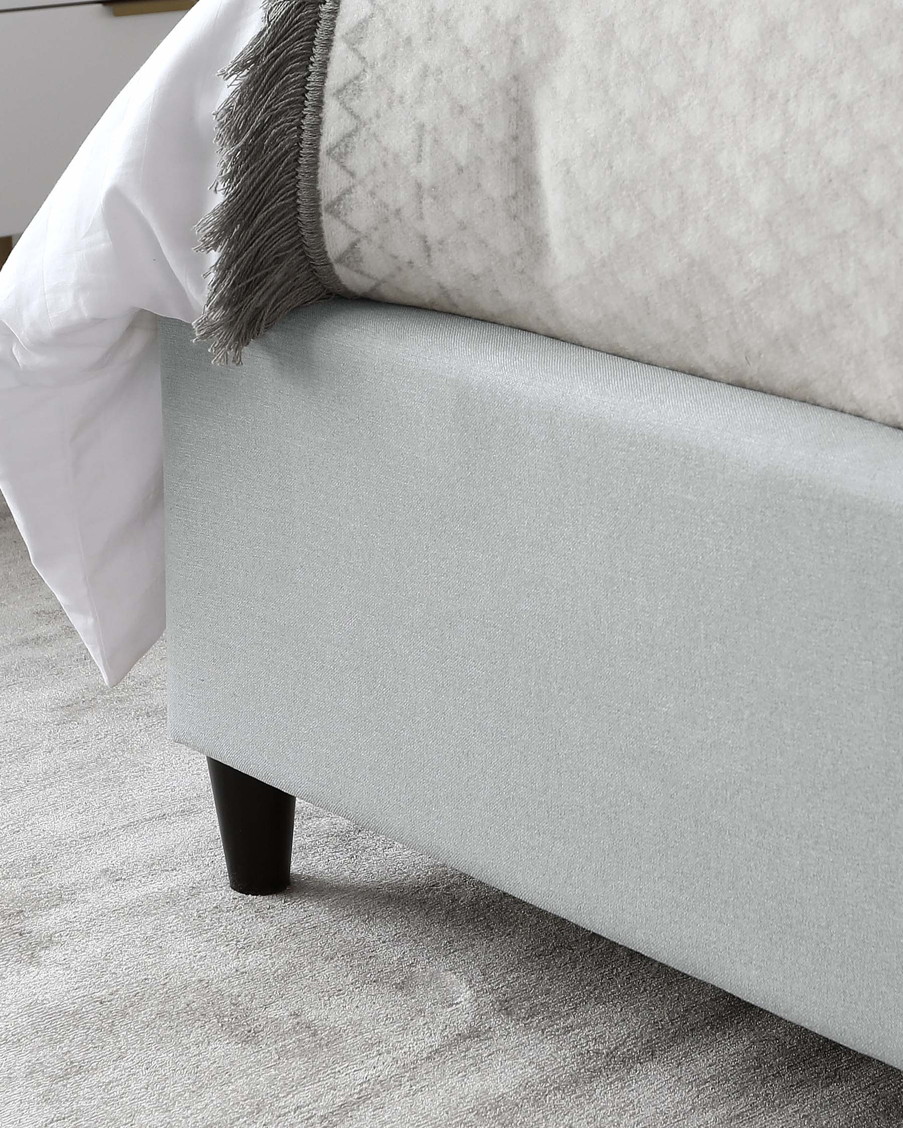 A close-up view of a section of a light grey upholstered bed frame with a simple and elegant design, featuring a smooth fabric finish and a clean, straight-line structure. The bed frame is supported by a dark, cylindrical wooden leg visible at the corner. A white bedsheet partially covers the bed, and a textured light grey throw blanket with tassel fringe lies atop the sheet, indicating a styled and cosy sleeping arrangement. The furniture rests on a soft, patterned area rug with varying grey tones.