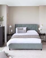 An elegant bedroom display featuring a plush, upholstered king-sized bed in a soft sage green with button tufted headboard detail. The bed is outfitted with crisp white bedding, a textural grey throw, and complementary decorative pillows. Flanking the bed are two black nightstands with a sleek, minimal design, each adorned with stylish table lamps and small decorative items. The scene exudes a serene, contemporary aesthetic.