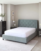 A contemporary styled bedroom featuring an upholstered grey platform bed with a button-tufted headboard, flanked by two black nightstands with silver handles. A matching black, tall dresser is also visible, paired with decorative items including a floor lamp, potted plant, and framed wall art. The room is completed with a soft grey area rug.