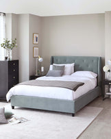 Elegant bedroom furniture set including an upholstered king-size bed with a tufted headboard in a soft grey tone, two matching nightstands with drawers, and a tall black dresser, all coordinated with a contemporary neutral colour scheme. The room is accessorized with decorative lamps, artwork, and a vase with greenery.