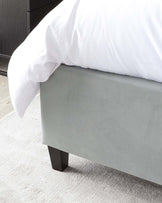 A contemporary light grey fabric upholstered bed frame with a clean-lined silhouette and solid black wooden legs, partially covered by a white duvet. The backdrop includes a dark-toned wooden nightstand or dresser, complementing the modern and minimalist aesthetic of the bedroom furniture.