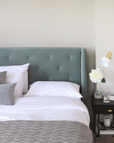 Elegant bedroom featuring a plush queen-sized bed with a tufted, seafoam green upholstered headboard and a coordinating patterned comforter. Beside the bed is a sleek, modern black nightstand with a simple white lamp, a clear vase with white flowers, and a glass of water on top, above which is a white, adjustable wall-mounted lamp.