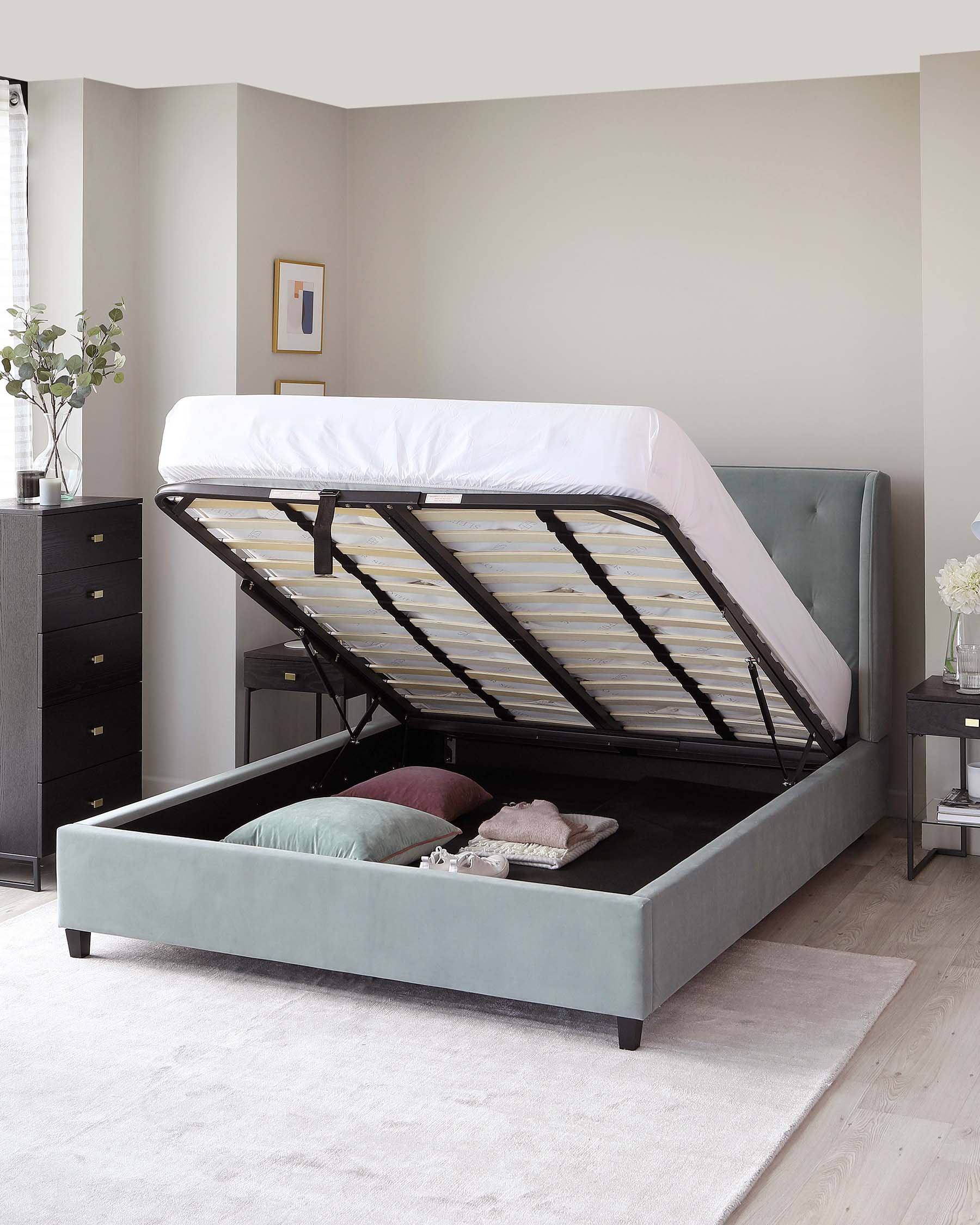 A contemporary upholstered storage bed in light grey fabric with the mattress lifted to showcase the ample under-bed storage space. The bed has a tufted headboard and is set on dark wooden feet. A black chest of drawers is visible to the left, coordinating with the bed's modern style.