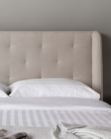 Upholstered modern bed headboard in a neutral colour fabric with button-tufted detail, accompanied by a white bedsheet and striped blanket at the foot of the bed.