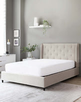 Contemporary beige upholstered bed frame with a tufted headboard, accompanied by a matching nightstand featuring a white table lamp, and a white floating shelf with decorative items above. The scene is complemented by a pale grey area rug under the bed.