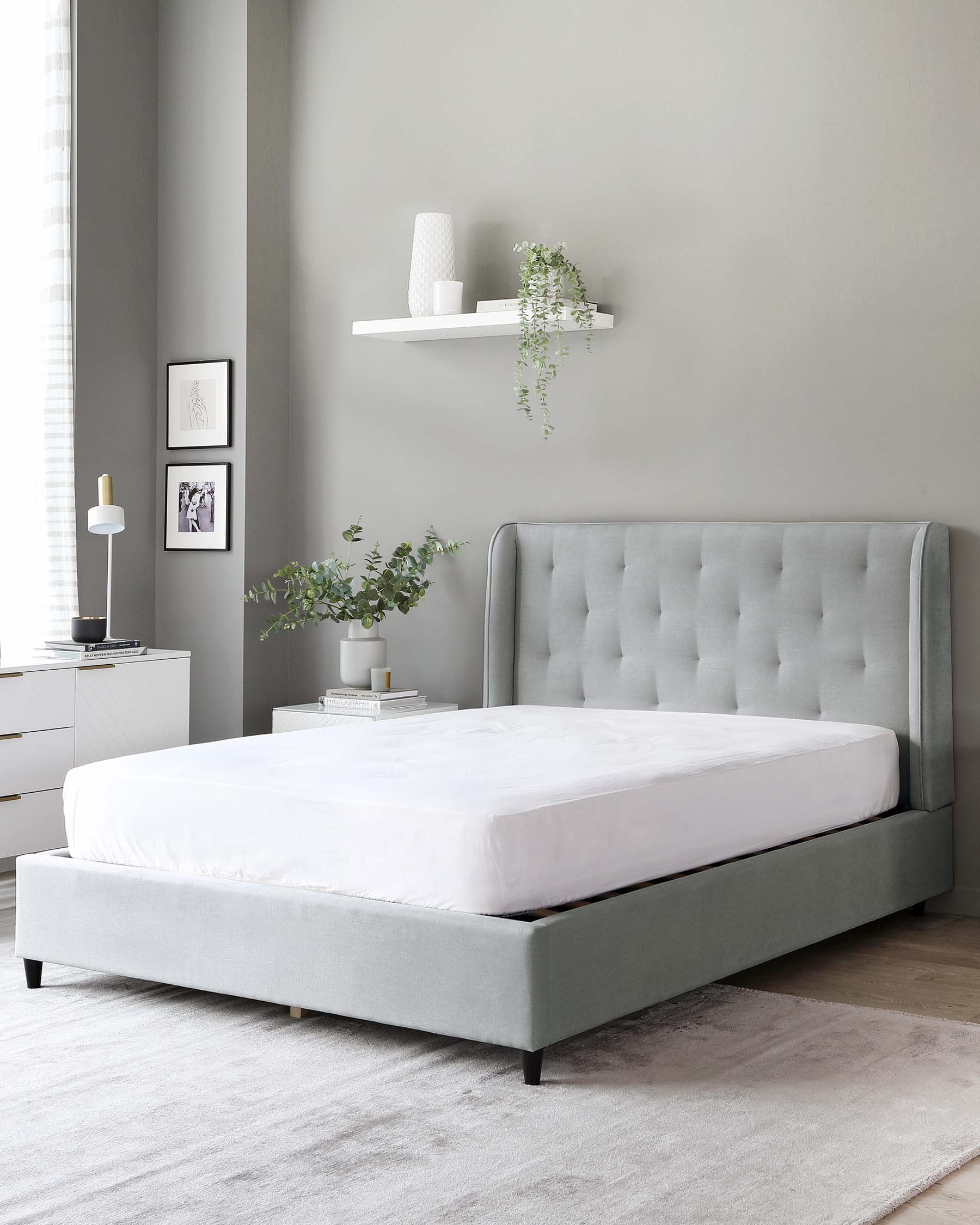 Modern bedroom with a grey upholstered platform bed featuring a tufted headboard, white bedding, and wooden legs. Beside it is a white two-drawer nightstand with a sleek design. Above the bed is a single white floating shelf with decor items.