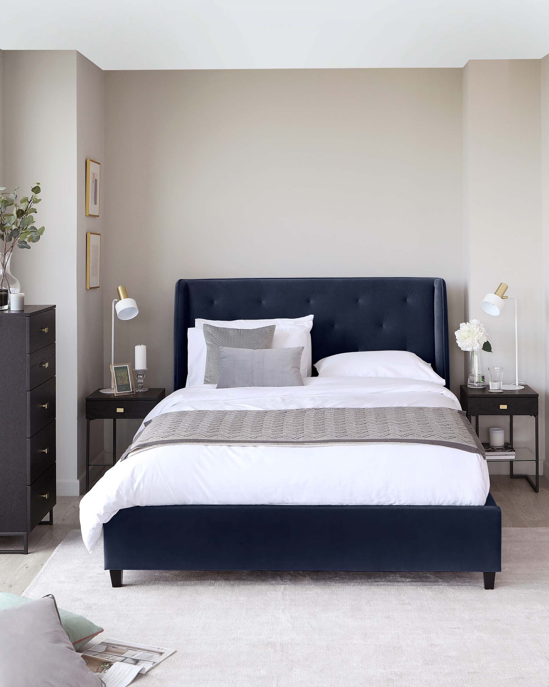 A contemporary bedroom featuring a dark navy upholstered bed with a tufted headboard, complemented by two matching nightstands with brass knobs and slender legs. Each nightstand hosts a white lamp and small decor items.