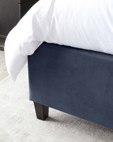 Navy blue upholstered bed bench with dark wooden legs, set against a light grey carpet and white bedding.