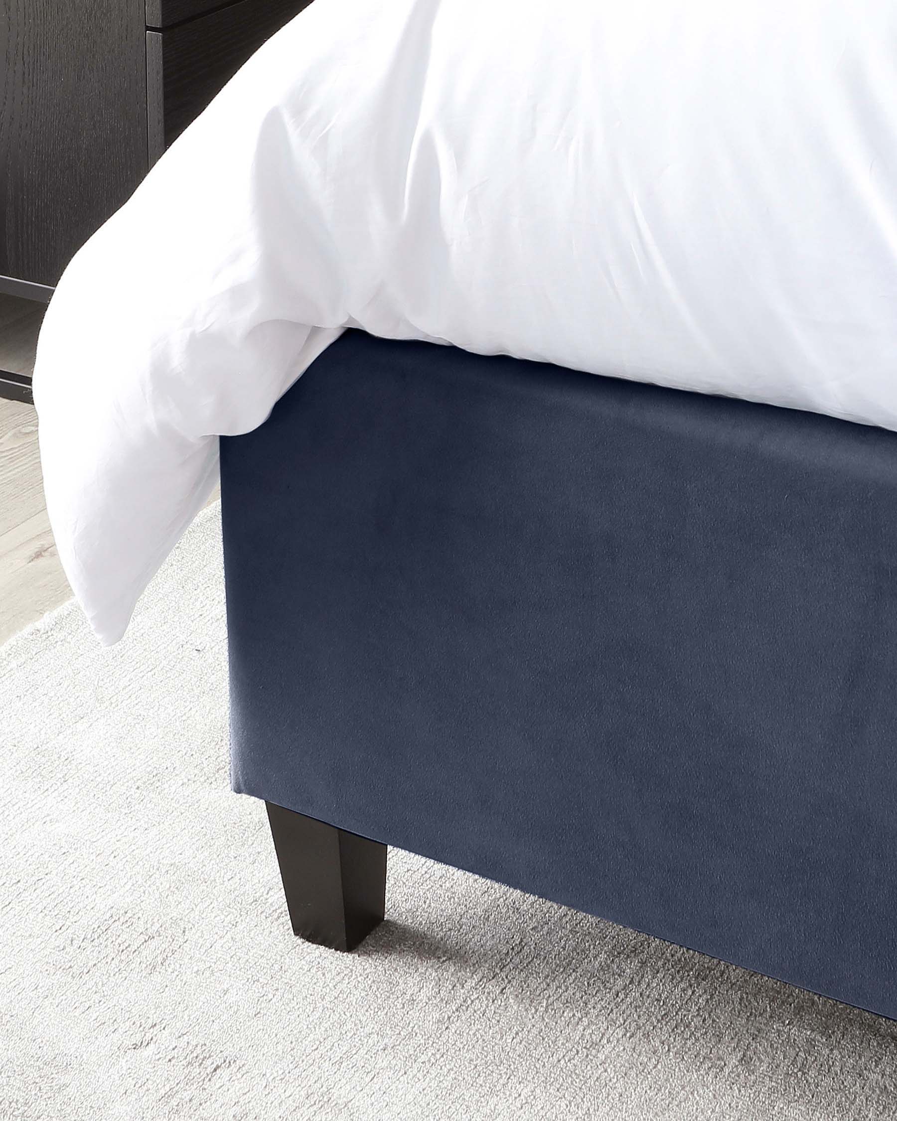 Navy blue upholstered bed bench with squared edges and black wooden legs, positioned at the foot of a bed with a white comforter, against a neutral carpeted floor and a dark bedside dresser in the background.