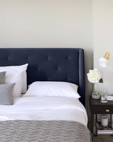 A contemporary bedroom featuring a navy blue upholstered headboard with tufted details, complemented by a simple black bedside table with a drawer and gold knob. A white and gold lamp sits atop the nightstand next to a clear vase with white flowers. The bed is dressed in crisp white linen with a grey textured throw at the foot and a mix of white and grey pillows.