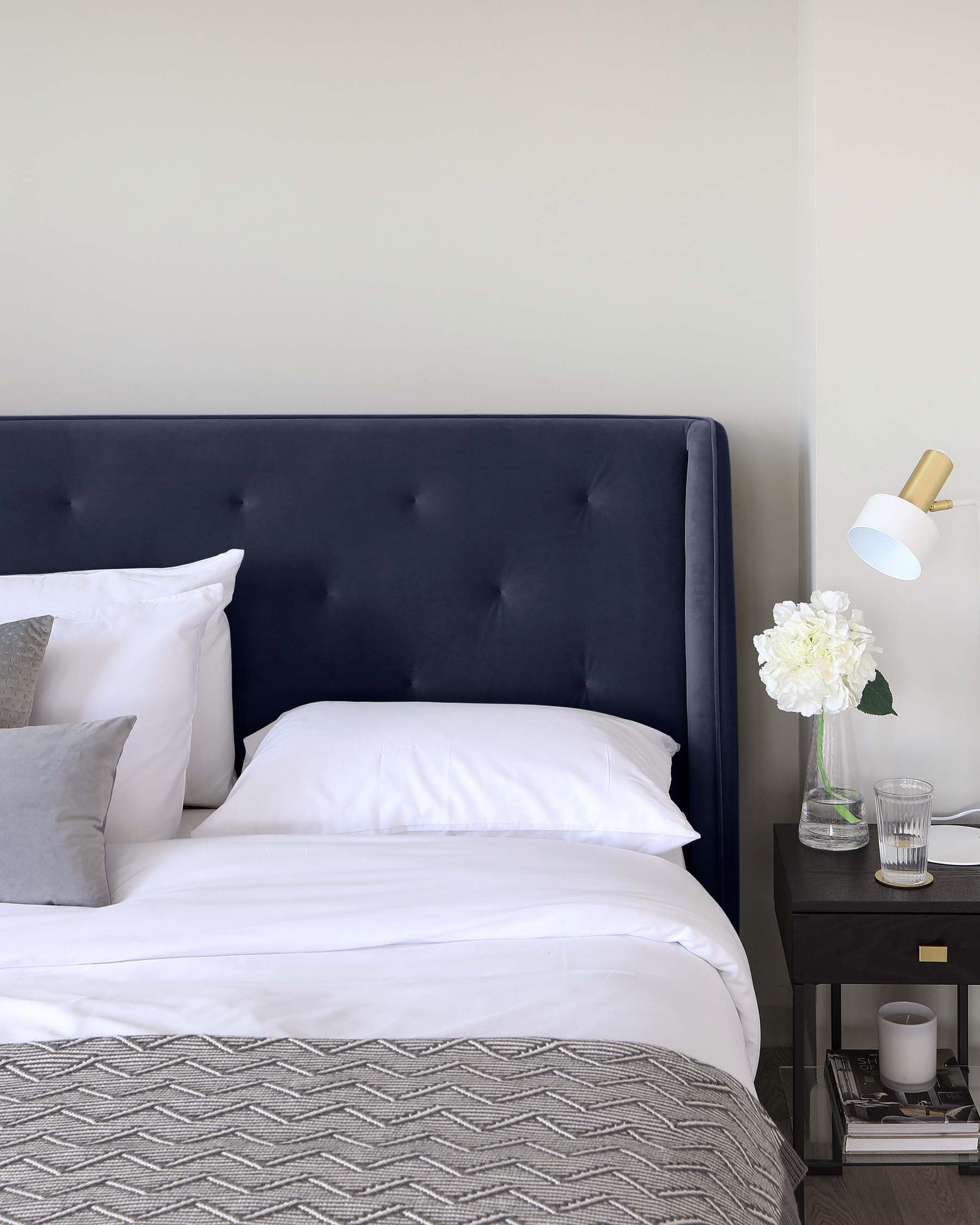 A contemporary bedroom featuring a dark blue upholstered bed with a tufted headboard, white and grey bedding, and a herringbone-patterned throw. Beside the bed is a black nightstand with a white and gold lamp, and decorative items including a vase with white flowers and a glass of water.