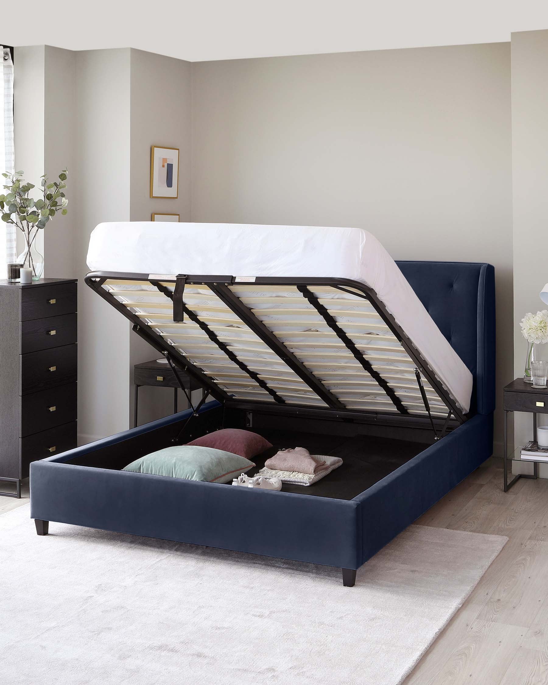 A contemporary navy blue upholstered storage bed with a lifted mattress platform revealing an ample storage space beneath. The bed features a sleek design with a simple headboard and solid wooden legs. To the right, there is a matching navy blue nightstand with a white floral arrangement on top and an open black drawer showcasing a neatly organized interior.