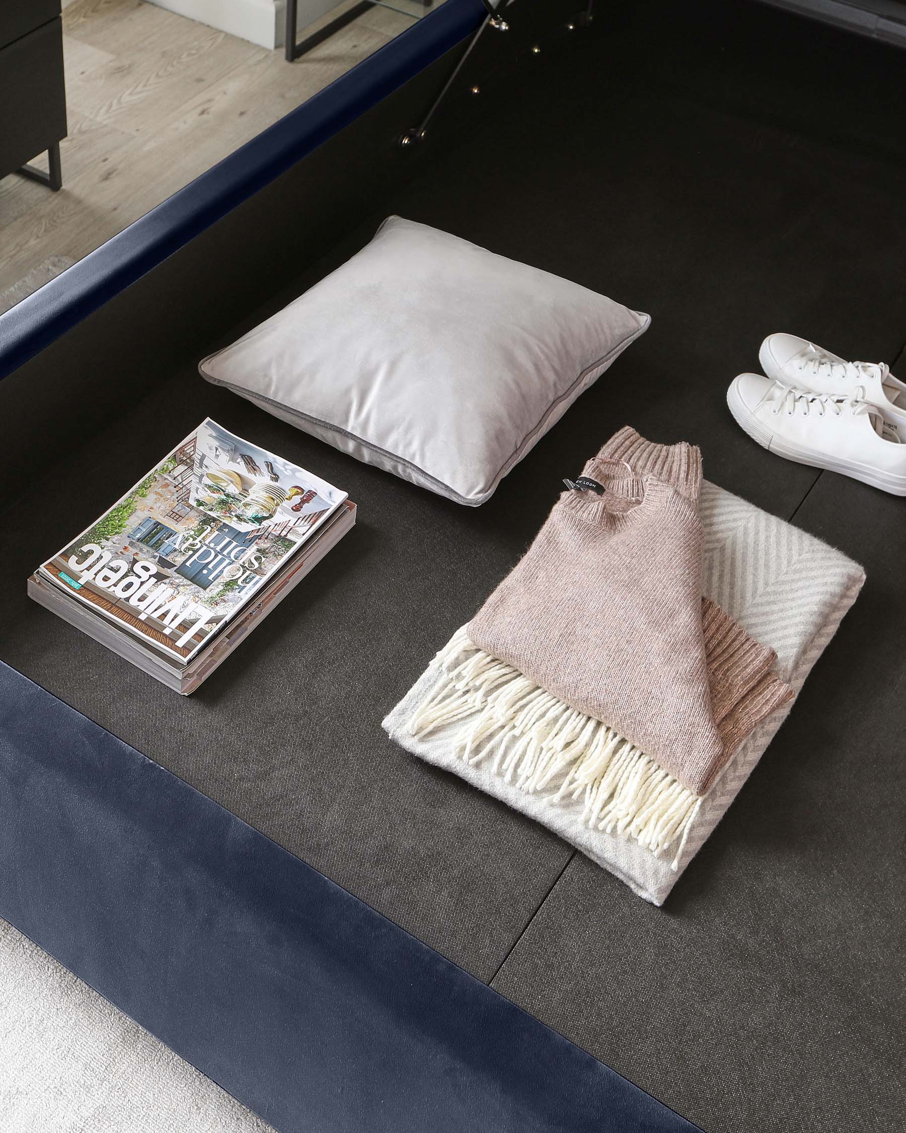 Modern low-profile rectangular ottoman in navy blue with a plush surface and a contrasting light grey magazine on top, accompanied by a silver decorative pillow and a folded beige sweater over a white fringed throw, set on a dark charcoal carpet.