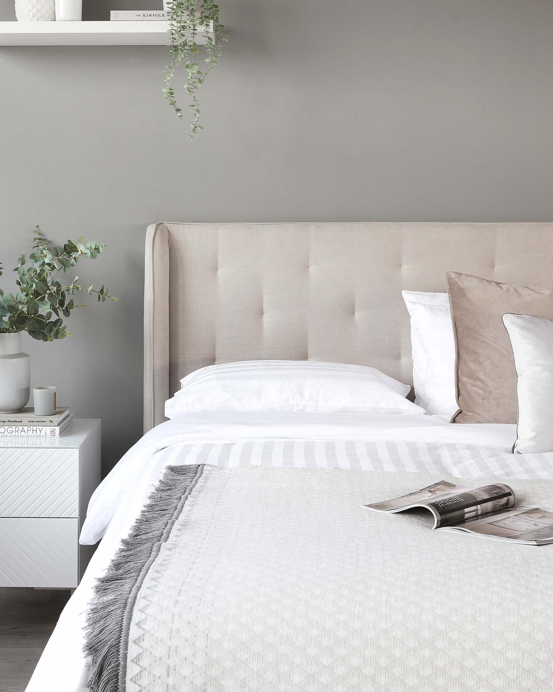 Elegant bedroom featuring a large upholstered tufted headboard in a neutral tone, coordinating with a sleek white bedside table adorned with decor and books. The bed is dressed in layered white and striped linens, accentuated by a textured throw and a mix of plush pillows.